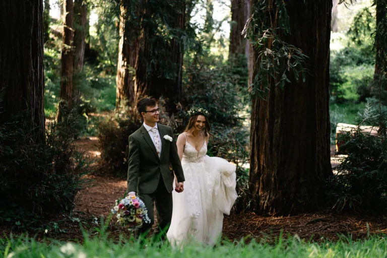 Elopement couple holding hands and walking through a forest trail surrounded by tall trees.
