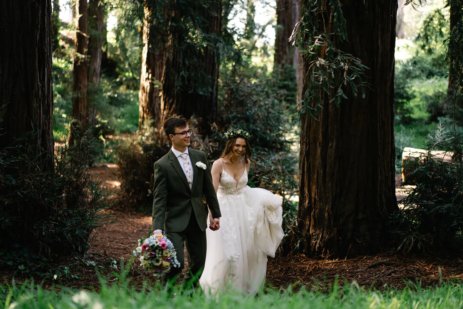 Elopement couple holding hands and walking through a forest trail surrounded by tall trees.