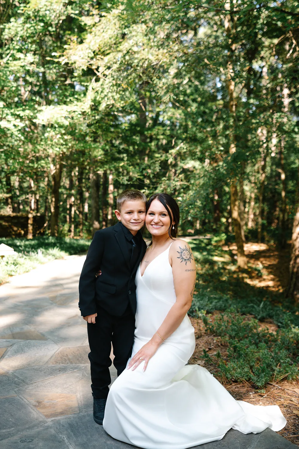 Bride standing with a young family member during a queer wedding at a wooded outdoor venue, smiling together in natural light.