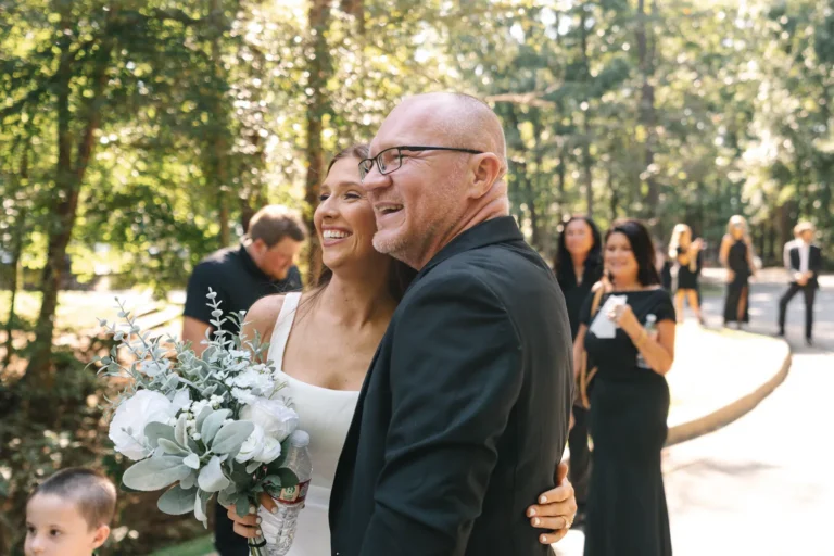 Bride smiling and hugging her dad after the ceremony, holding a bouquet while guests gather in the background outdoors.