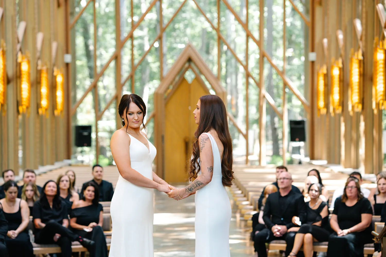 Two brides hold hands during their wedding ceremony inside Anthony Chapel, standing at the altar with guests seated behind them.
