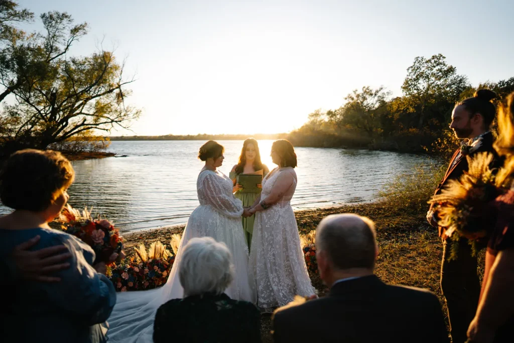 Two brides holding hands during an intimate lakeside wedding ceremony at sunset with loved ones gathered nearby