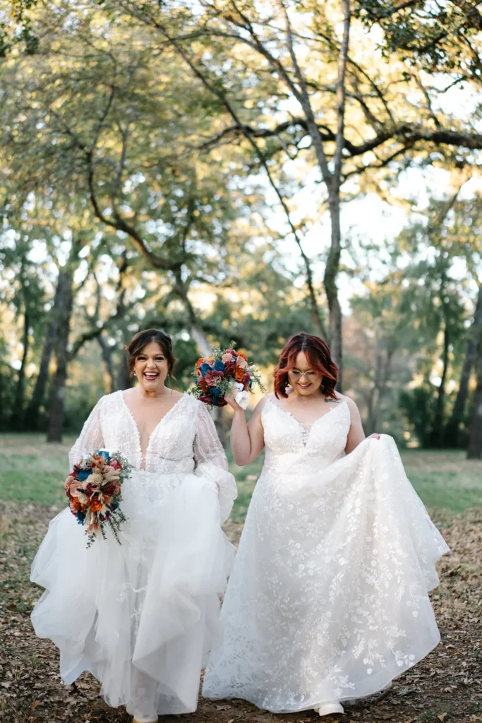 Two brides walking hand in hand through a wooded area before their wedding ceremony, holding bouquets and smiling