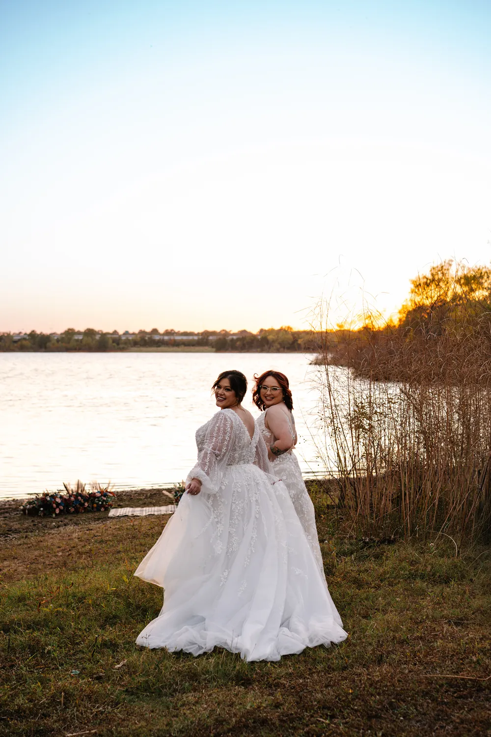 Two brides standing together by a lake at sunset, smiling back at the camera during an intimate wedding