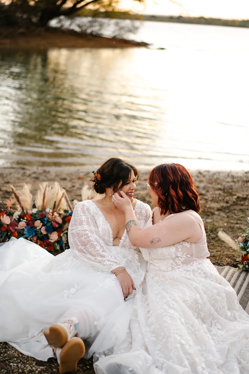 Two brides sitting together by a lake during an intimate wedding ceremony, smiling as one gently touches the other’s face.