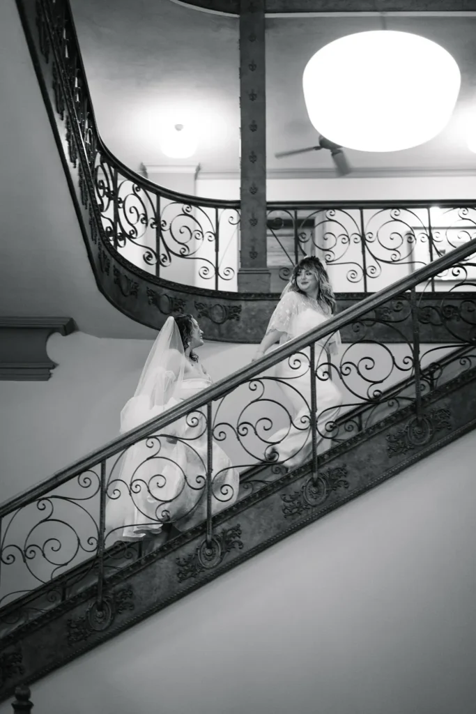 Two brides in wedding dresses walk up an ornate staircase inside the Fort Worth courthouse during their queer LGBTQ elopement.