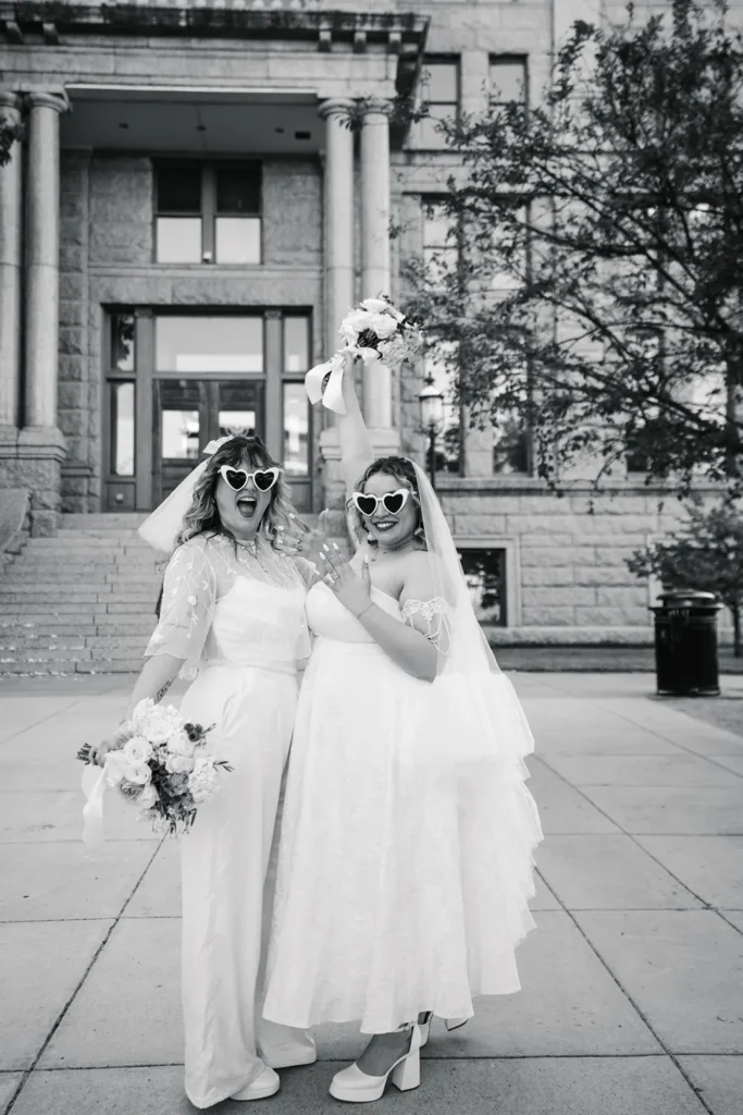 Two brides wearing heart-shaped sunglasses celebrate their queer LGBTQ elopement outside the Fort Worth courthouse.