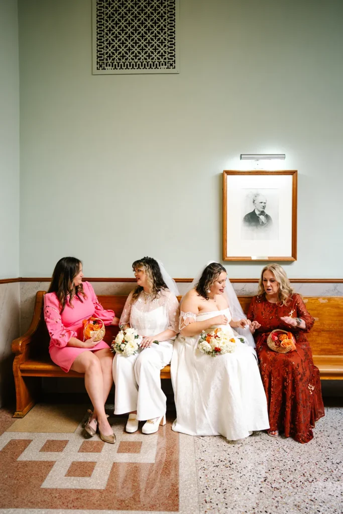 Two brides sit on a wooden bench inside the Fort Worth courthouse during a queer LGBTQ elopement, holding bouquets and talking with family members.